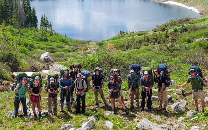 A group of students pose for a photo in front of a small body of water that is surrounded by green grass.